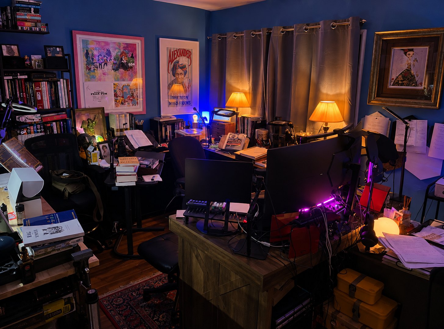 The room at night: blue walls, packed bookshelves, framed art including an Alexander the Man Who Knows poster, two desks with monitors and lit lamps, papers and cases stacked around a patterned rug.