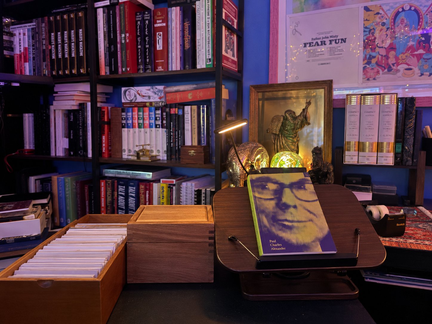 An open card box full of index cards and a second closed box on the desk, beside a book propped open on a stand under a task lamp, with packed bookshelves behind.